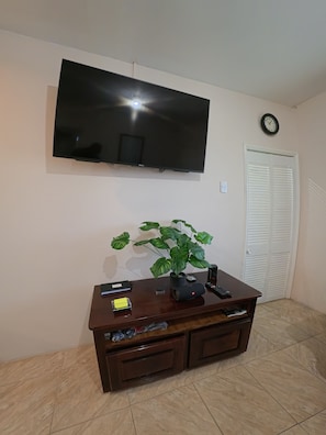 A living room with a flat-screen TV mounted on the wall, a wooden entertainment center with a plant, and a white door with louvered shutters. - Sweet Escape Welcoming  2-bedroom apartment  Kelly Village with AC comfort (Kelly Village)