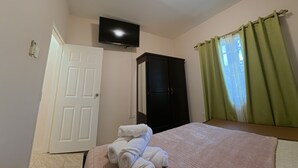 Ein Schlafzimmer mit einem Bett, einem an der Wand montierten Fernseher, einem Kleiderschrank und einem Fenster mit Vorhängen.