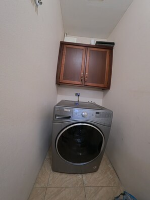 A compact laundry area with a front-loading washing machine, wooden cabinets above, and tiled flooring. - Sweet Escape Welcoming  2-bedroom apartment  Kelly Village with AC comfort (Kelly Village)