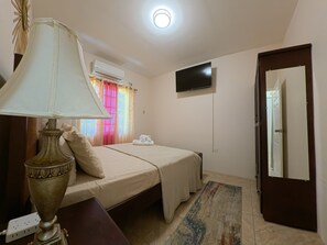 A hotel room with a bed, a bedside lamp, an air conditioning unit, a television, and a door. - Sweet Escape Welcoming  2-bedroom apartment  Kelly Village with AC comfort (Kelly Village)