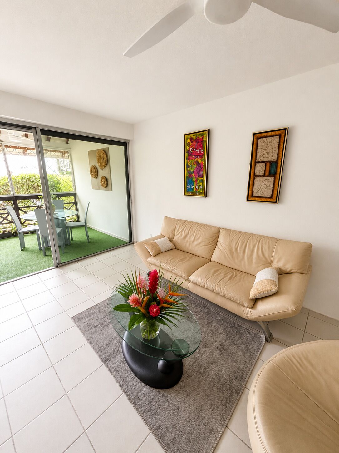 A living room with a beige sofa, a glass coffee table with a floral centerpiece, and a sliding glass door leading to an outdoor area with a table and chairs.