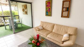 A living room with a beige sofa, a glass coffee table with a floral centerpiece, and a sliding glass door leading to an outdoor area with a table and chairs.