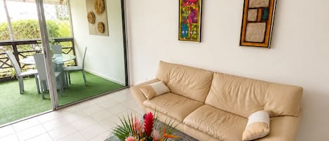 A living room with a beige sofa, a glass coffee table with a floral centerpiece, and a sliding glass door leading to an outdoor area with a table and chairs.