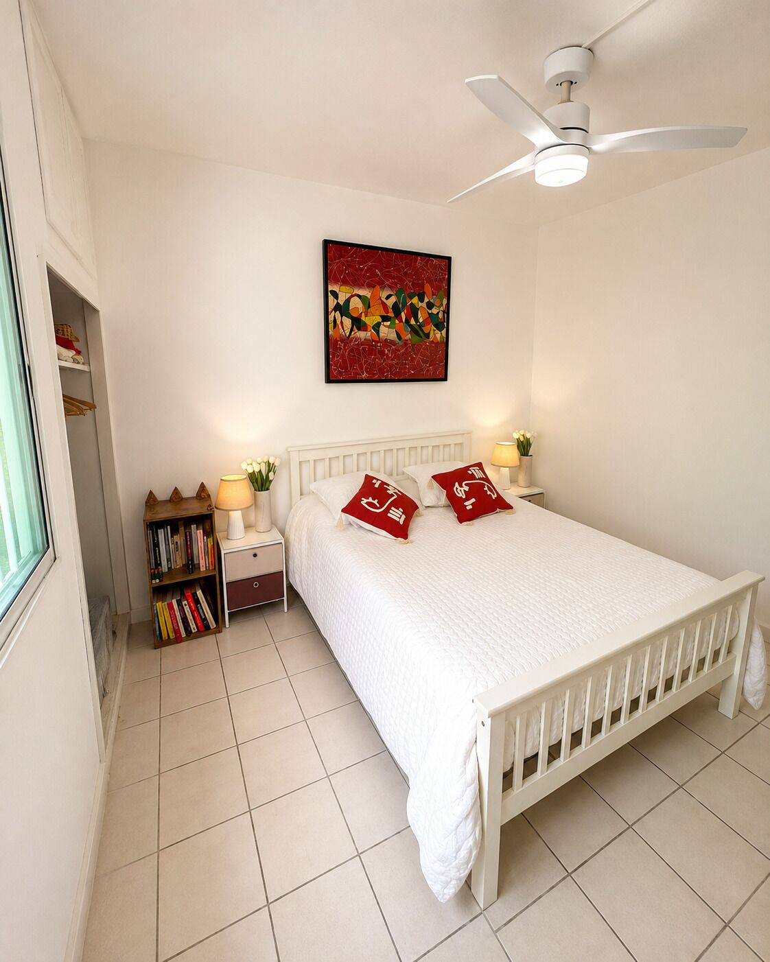 A bedroom with a white bed, a ceiling fan, a bedside table with a lamp, a bookshelf, and a wall-mounted artwork.