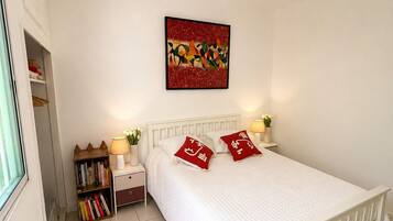 A bedroom with a white bed, a ceiling fan, a bedside table with a lamp, a bookshelf, and a wall-mounted artwork.