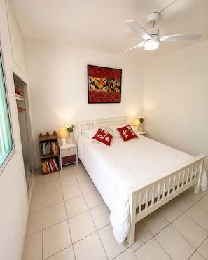 Un dormitorio con cama blanca, ventilador de techo, mesita de noche con lámpara, estantería y una obra de arte colgada en la pared.