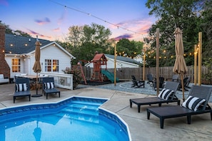 A backyard with a swimming pool, lounge chairs, a wooden play structure, and string lights. - 4BR Backyard Oasis: Pool, Hot Tub & Fire Pit (Charlotte)