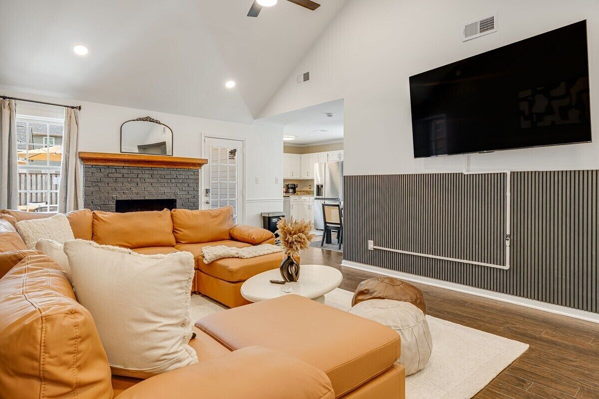 A living room with a sectional sofa, a fireplace, a flat-screen TV, and a kitchen in the background.
