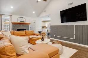 A living room with a sectional sofa, a fireplace, a flat-screen TV, and a kitchen in the background. - 4BR Backyard Oasis: Pool, Hot Tub & Fire Pit (Charlotte)