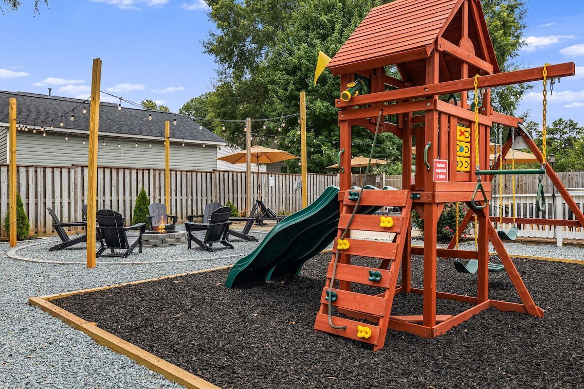 A playground with a slide and climbing structure, surrounded by a wooden fence, outdoor seating, and a house in the background.