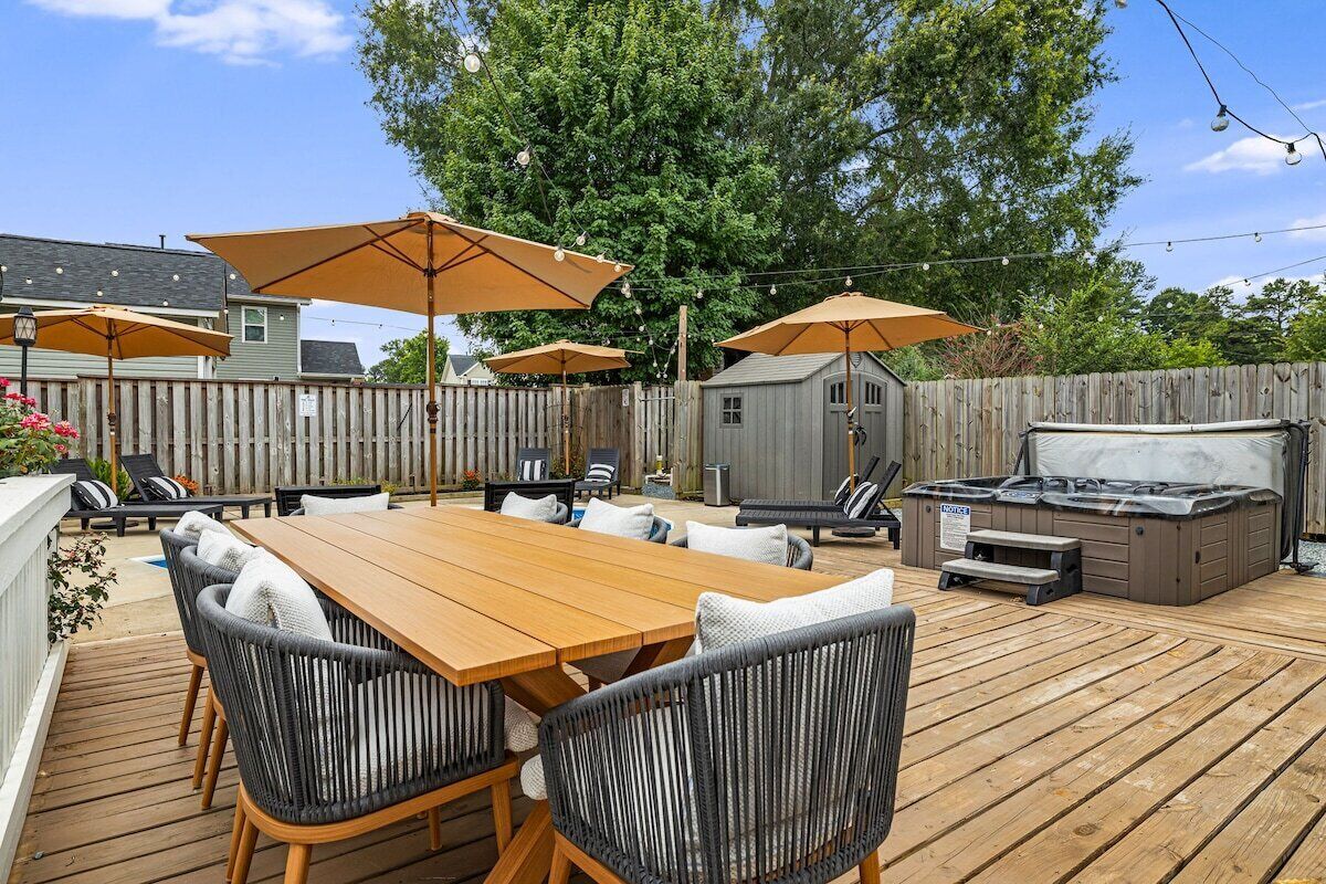 An outdoor patio with a wooden table and chairs, surrounded by umbrellas, and a hot tub.