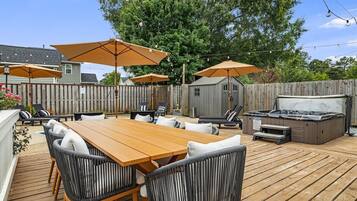 An outdoor patio with a wooden table and chairs, surrounded by umbrellas, and a hot tub.