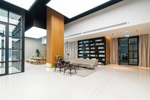 Eine geräumige Lobby mit hoher Decke, einer großen Bücherwand, einer Sitzecke mit Sofa und Sesseln sowie einem Glastür-Eingang.