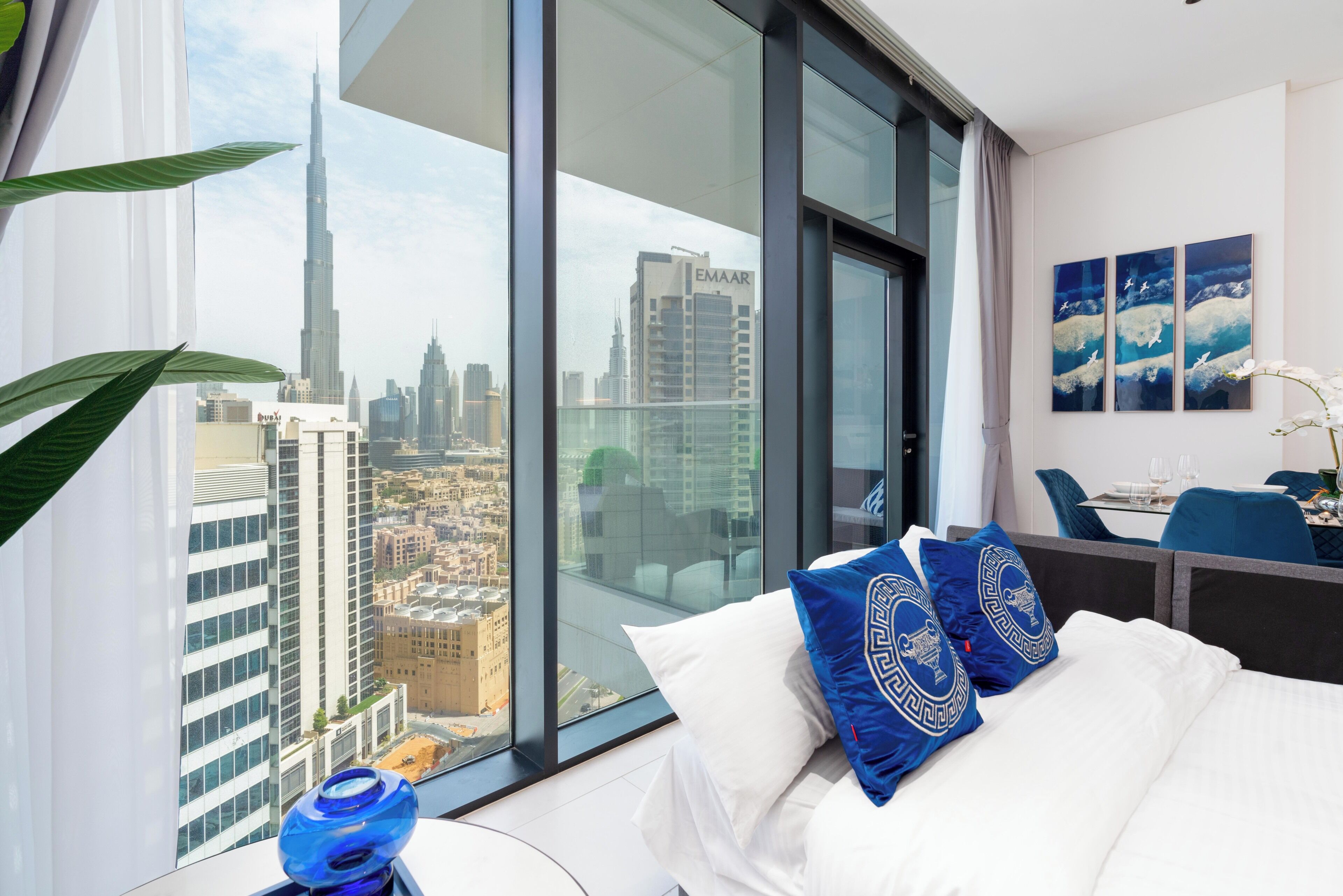Ein Hotelzimmer mit großem Fenster und Blick auf die Stadt, ein Bett mit weißer Bettwäsche und blauen Zierkissen, ein kleiner Tisch mit Glasplatte sowie ein Sessel.