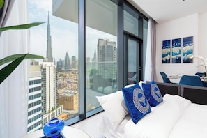A hotel room with a large window offering a city view, a bed with white bedding and blue decorative pillows, a small table with a glass top, and a chair. - bnbme - Marquise Square Tower - 2BR (Dubai)