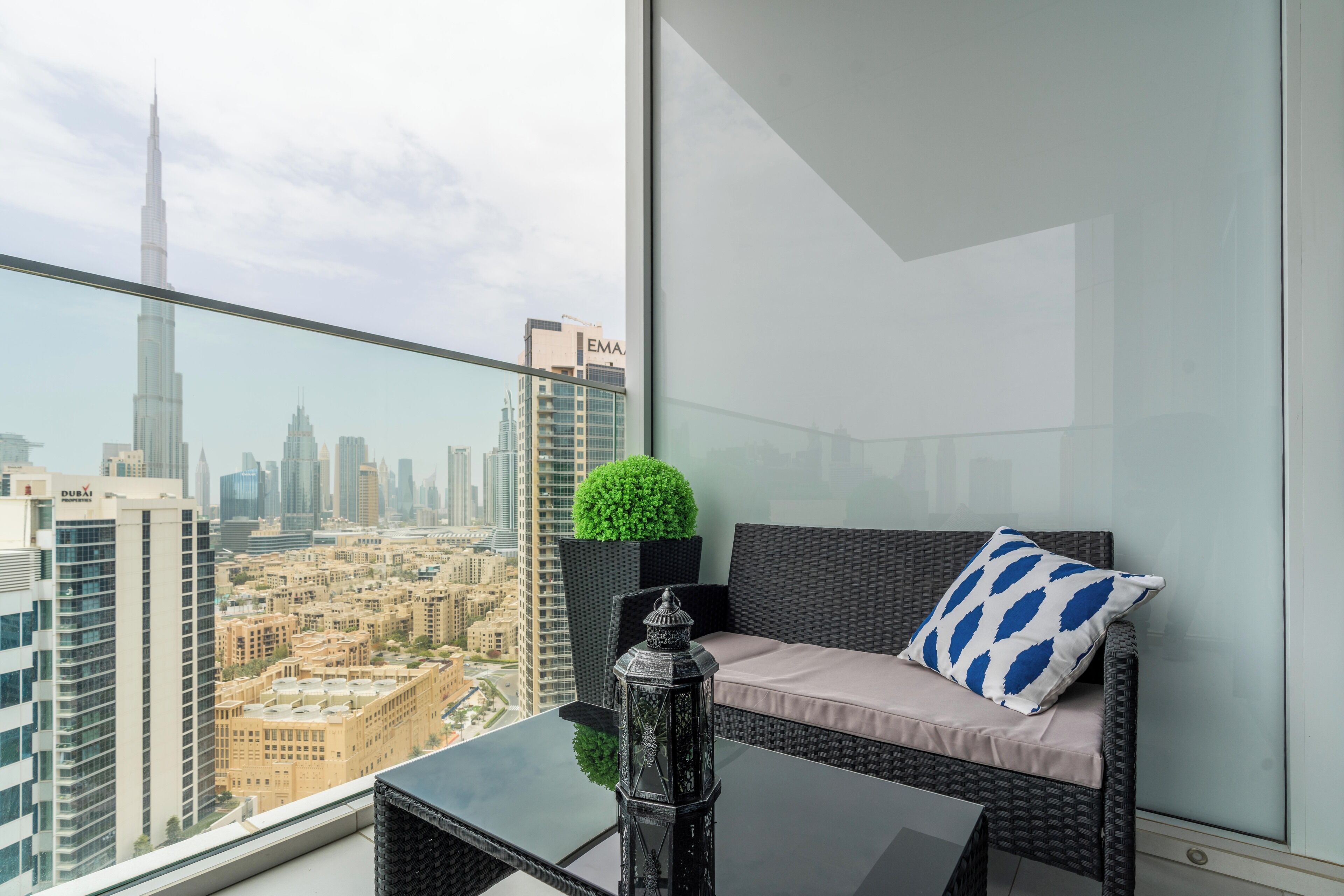 A balcony with a view of the Dubai skyline, including the Burj Khalifa, a wicker sofa with a cushion, a small potted plant, and a decorative item on a glass table.