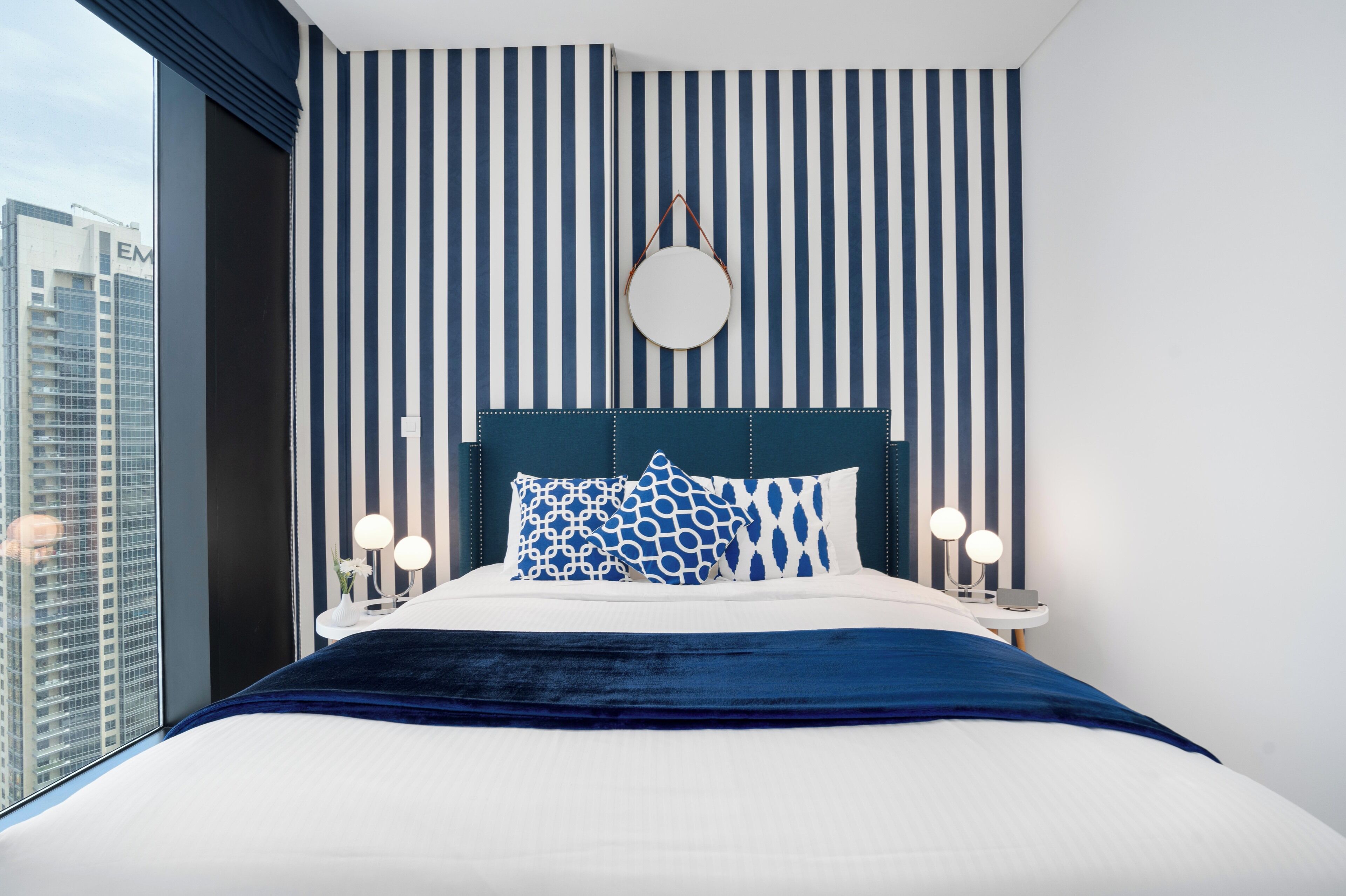A modern bedroom with a striped wall, a round mirror, and a bed with patterned pillows.
