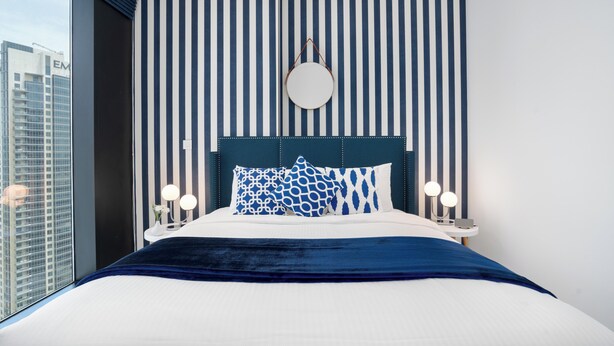 Un dormitorio moderno con pared a rayas, un espejo redondo y una cama con almohadones estampados.