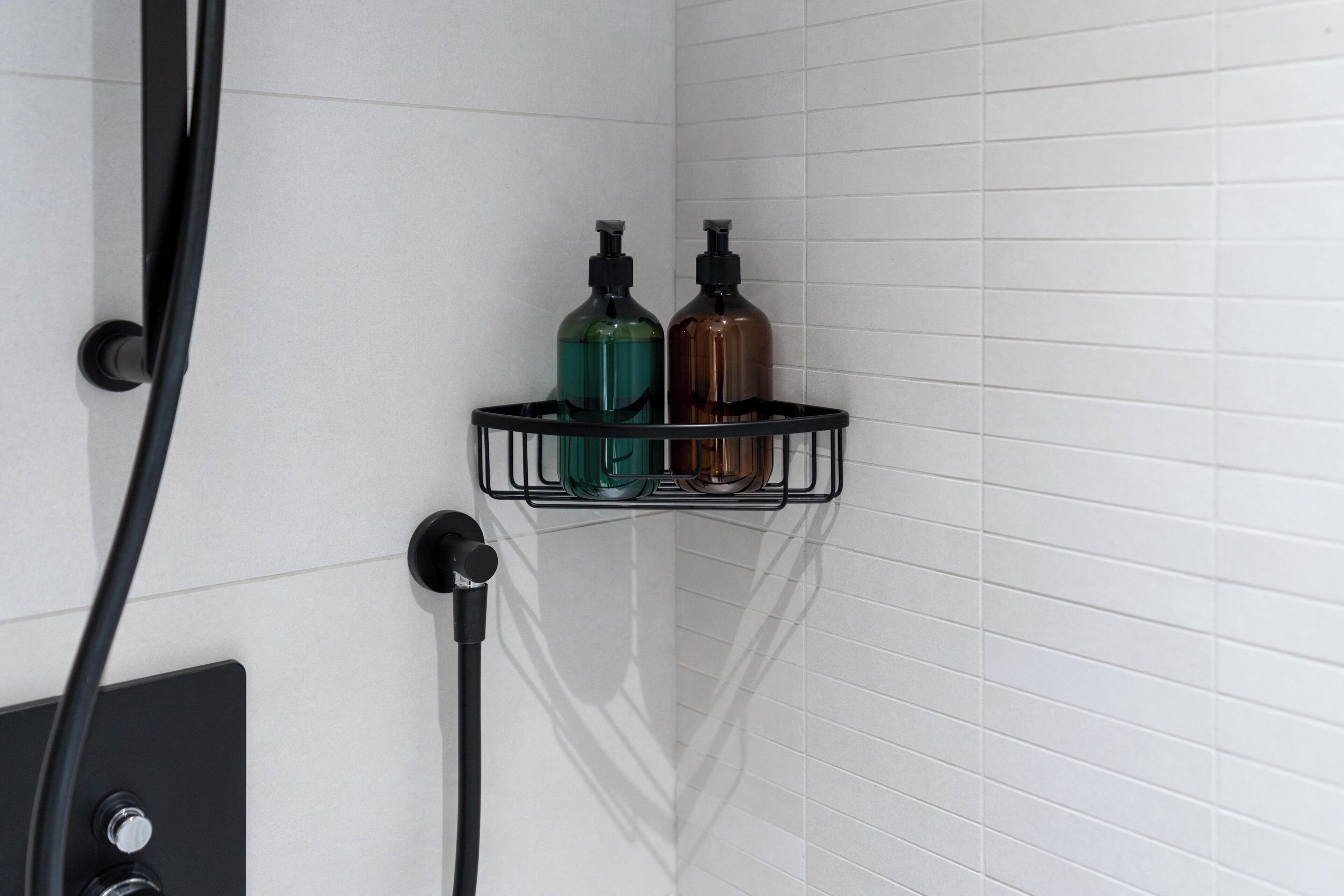 A corner of a bathroom with a black towel rack holding two bottles, a black faucet, and a wall-mounted light switch.