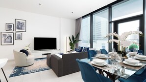 Una sala de estar moderna con televisor de pantalla plana, un sofá, una zona de comedor con sillas azules y un ventanal con vistas a la ciudad.
