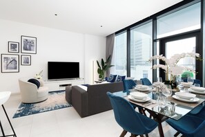 A modern living room with a flat-screen TV, a sofa, a dining area with blue chairs, and a large window offering a city view. - bnbme - Marquise Square Tower - 2BR (Dubai)
