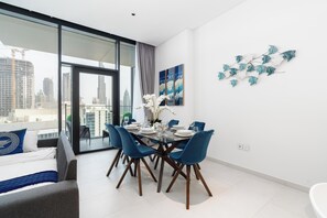 A modern dining area with a glass table, blue chairs, and a city view. - bnbme - Marquise Square Tower - 2BR (Dubai)