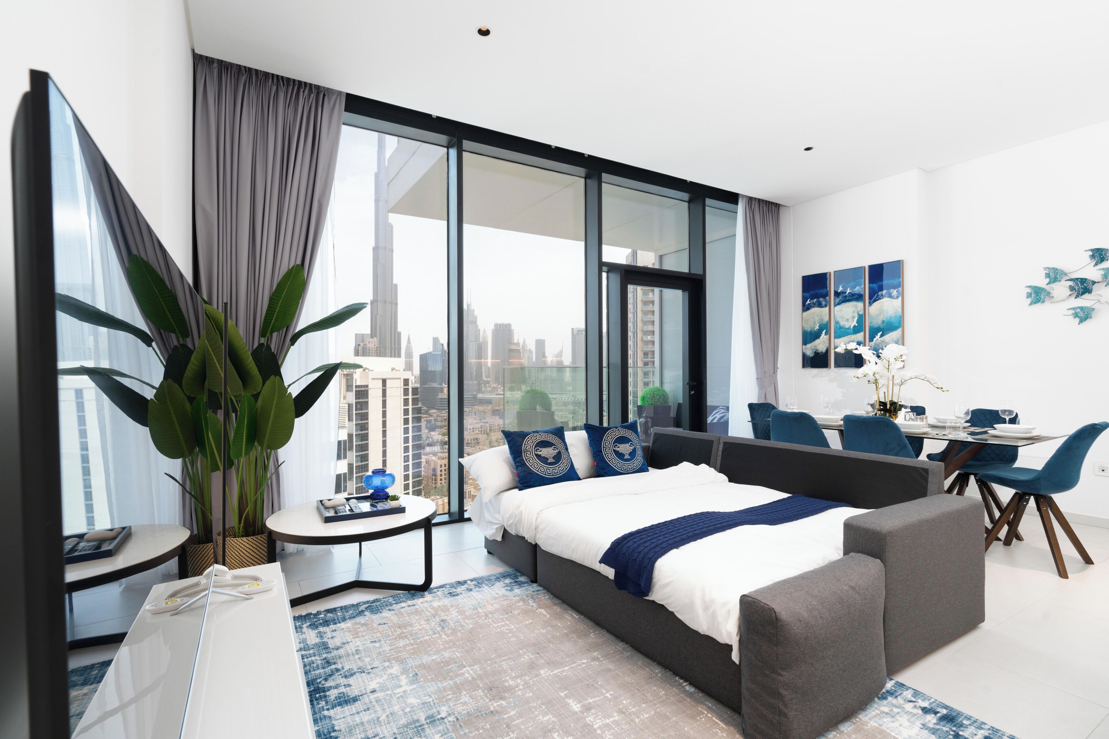 A modern hotel room with a large bed, a seating area, a dining table, and a view of the cityscape.