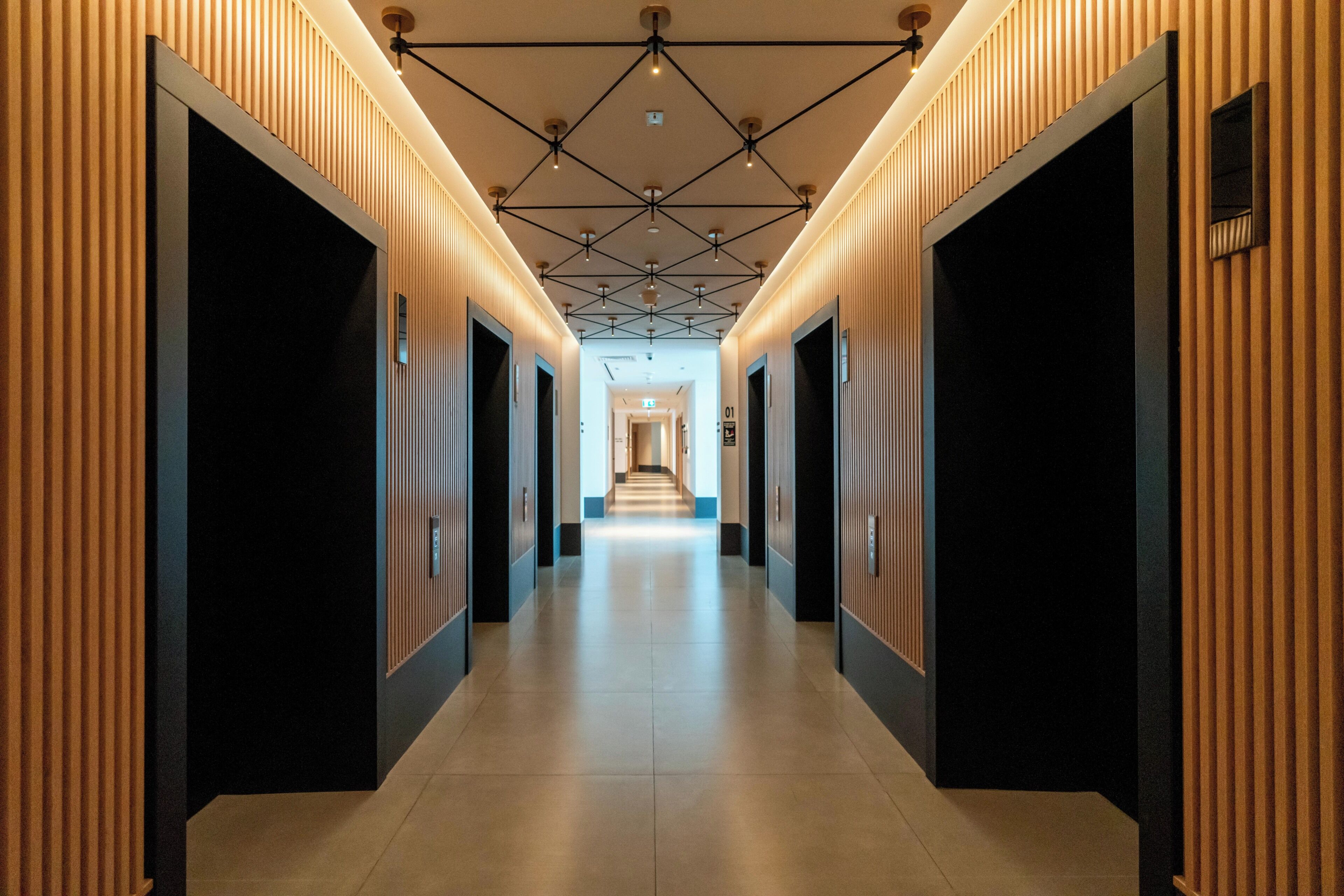 Un couloir moderne doté d’un plafond au motif géométrique, flanqué de panneaux de bois verticaux et de portes noires.