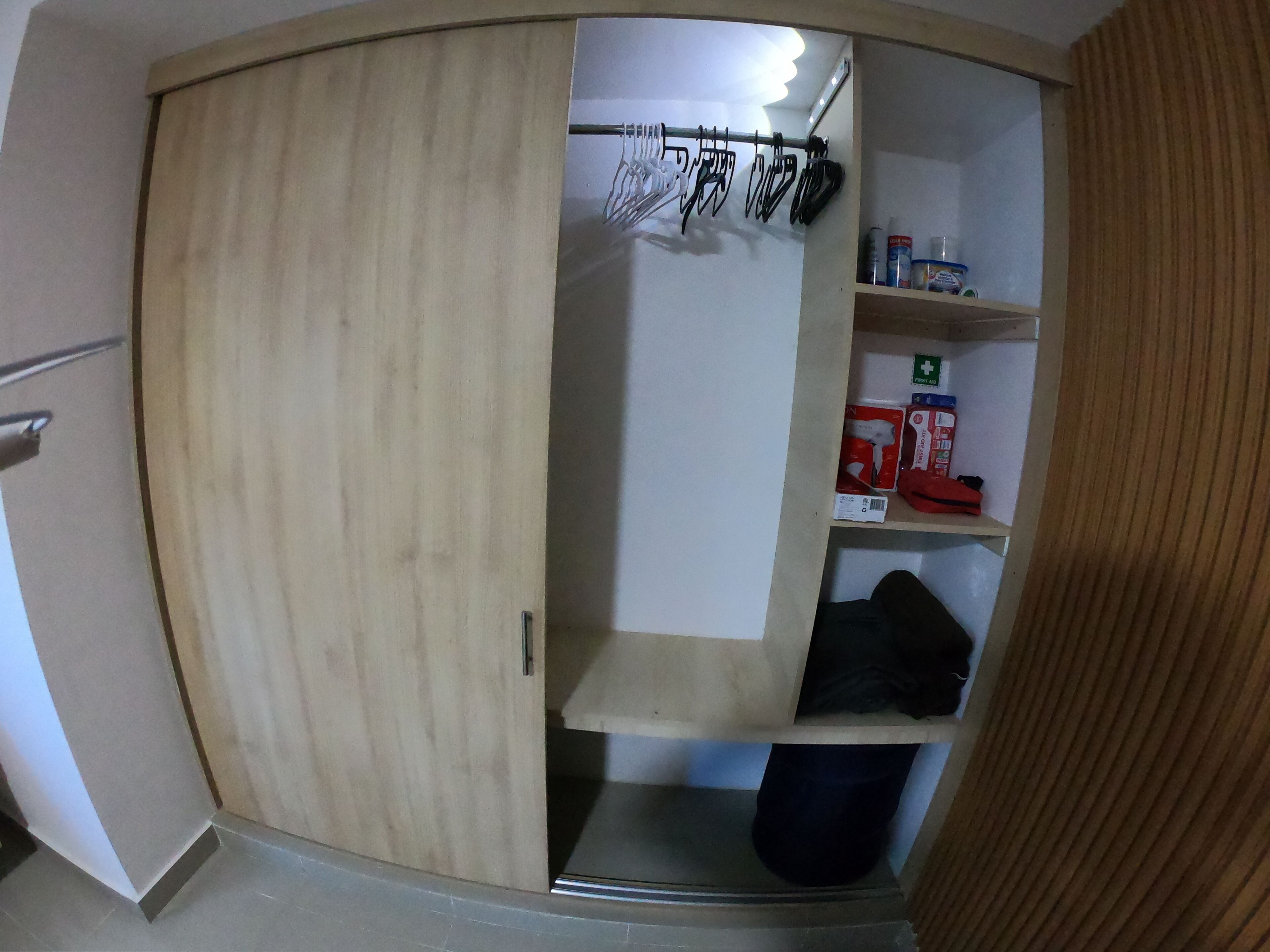 A room with a wooden wardrobe, a shelf with first aid supplies, and a hanging rack with utensils.