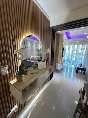 A modern interior with a wooden accent wall, a built-in desk with a mirror, and a view of a dining area with natural light. - Torre Samantha (Santo Domingo)