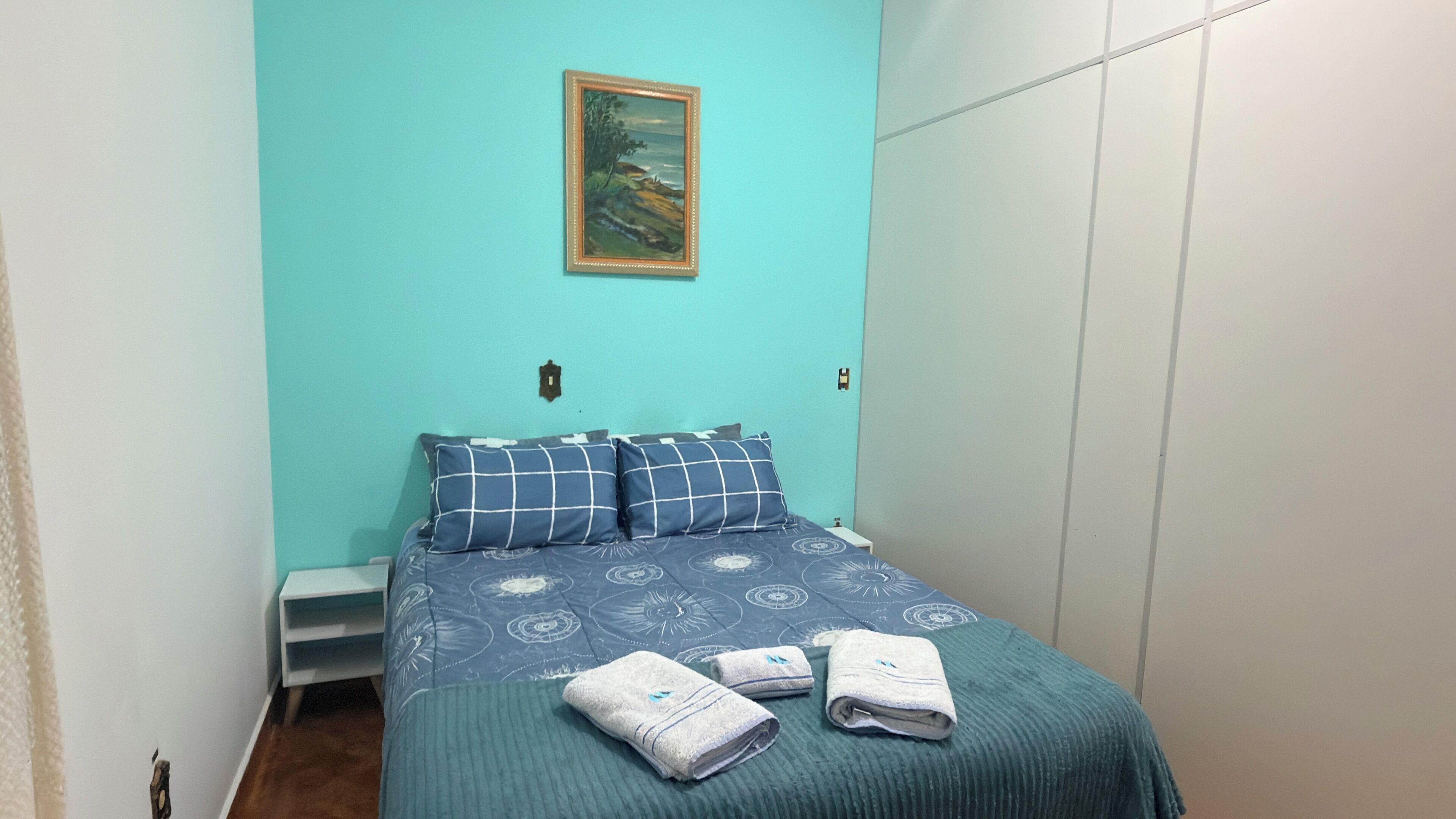 Quarto com cama, almofadas, um quadro e toalhas.