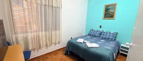 A bedroom with a bed, a window with curtains, a painting on the wall, and a tiled floor.