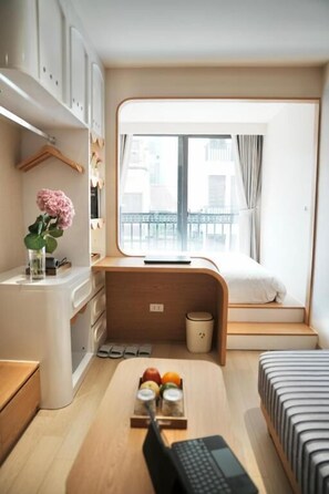 A compact bedroom with a bed, a small desk, a window with a view of buildings, and a trash bin. - D'la Lento Homestay (Hanoi)
