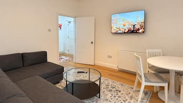 Una sala de estar con un sofá, una mesa de centro de vidrio, una mesa de comedor con sillas, un televisor montado en la pared y una puerta que da a otra habitación.