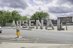 A road with large concrete letters spelling 'ARSENAL' and a yellow directional sign.