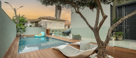 A swimming pool with a wooden deck, lounge chairs, and a tree.
