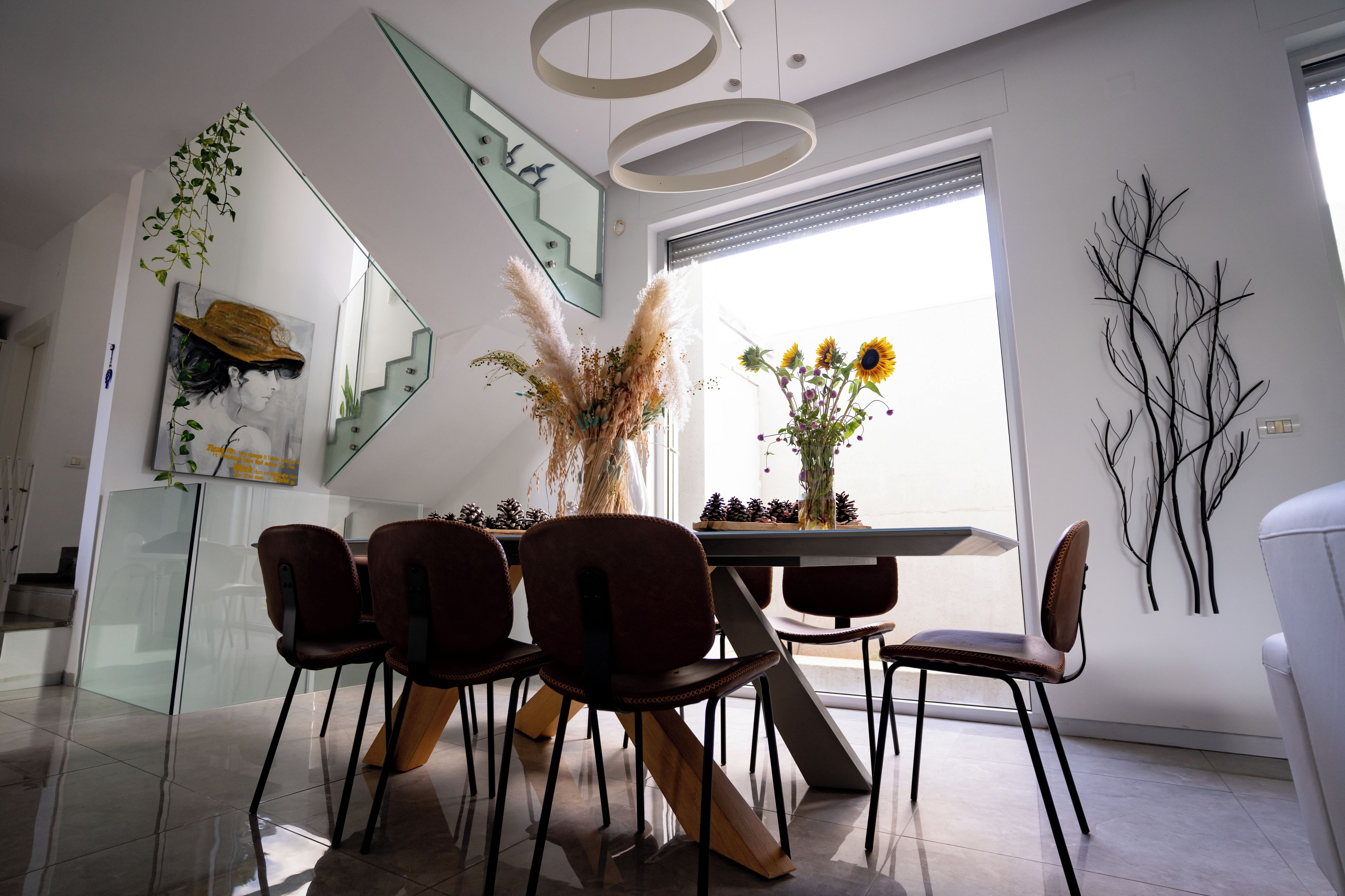 A modern dining area with a glass staircase, a dining table with chairs, and a vase of flowers.
