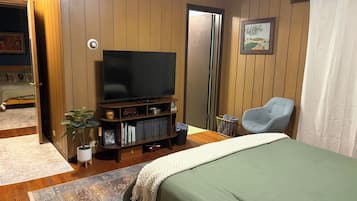 Un dormitorio con una cama, un televisor sobre un soporte de madera, una silla y una puerta que da a otra habitación.