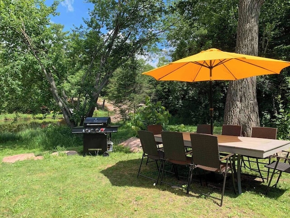 An outdoor dining area with a table and chairs, a grill, and a large umbrella. - Beautiful River-front Log Cottage: Glass Room & Hot Tub (Kawartha Lakes)