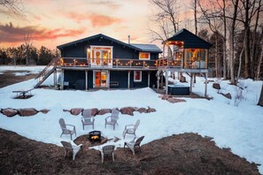 A two-story cabin with a deck, outdoor seating, and a fire pit. - Beautiful River-front Log Cottage: Glass Room, Hot Tub & Sauna (Kawartha Lakes)