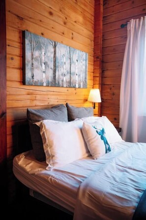A wooden cabin room with a bed, pillows, a lamp, and a painting on the wall.