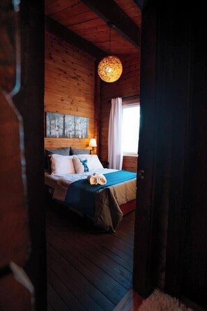 A wooden cabin room with a bed, a hanging light fixture, and a window with curtains.