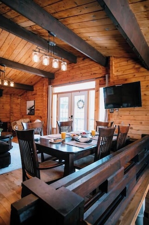 A wooden interior with a dining table, chairs, a bench, a television, and a window with a wreath.