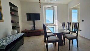 Un comedor con mesa y sillas de vidrio, un televisor sobre un mueble bajo, y vistas al exterior a través de un ventanal grande.