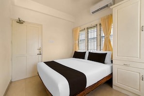 A hotel room with a bed, a wardrobe, a window with curtains, and an air conditioning unit. - Classic The Daffodils (Thiruvananthapuram)