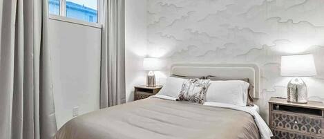 Una habitación de hotel con una cama, dos mesitas de noche, un ventanal con cortinas y una pared con diseño texturizado.