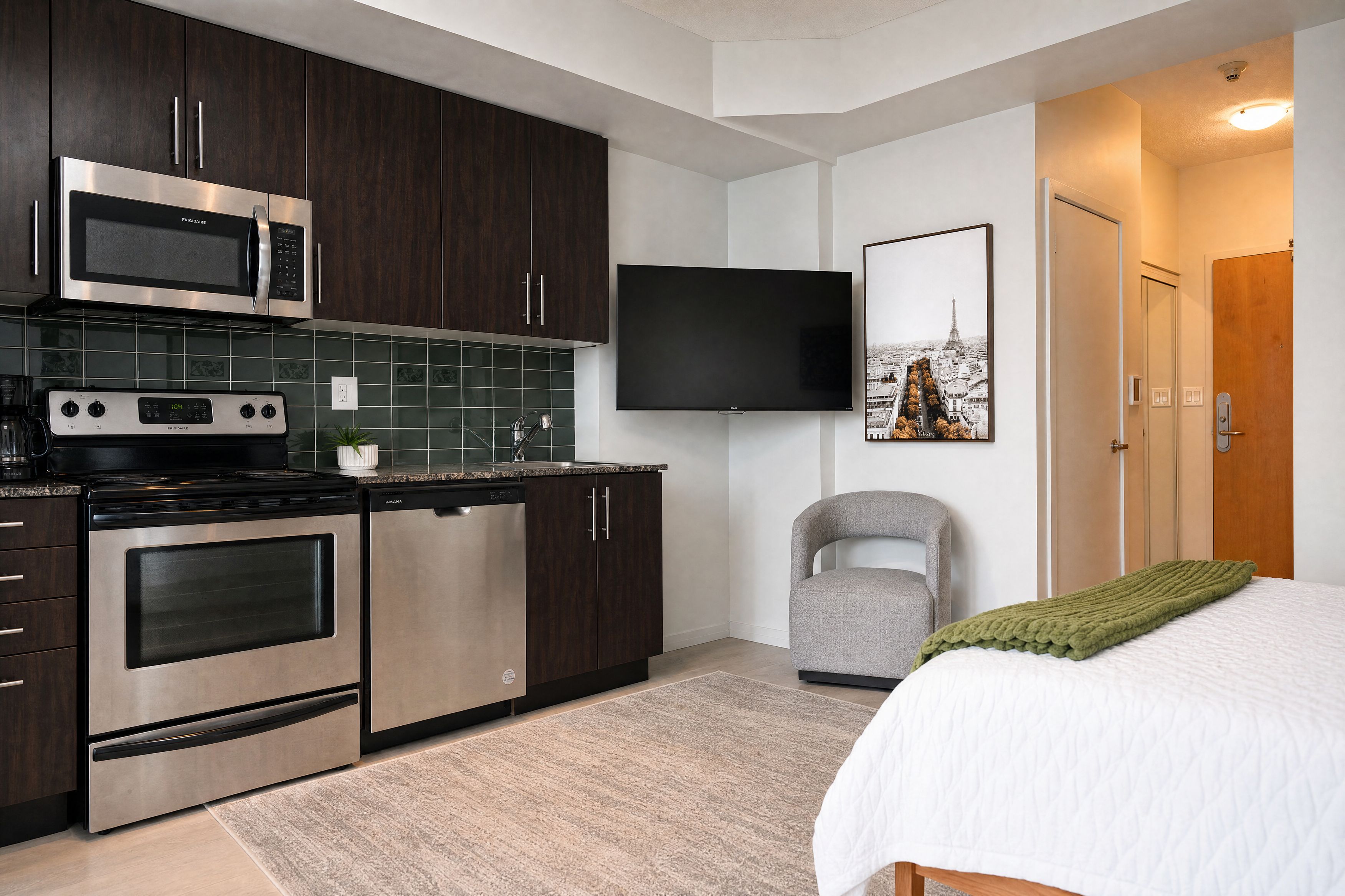 A modern kitchen with stainless steel appliances, a flat-screen TV, and a bed with a green blanket.