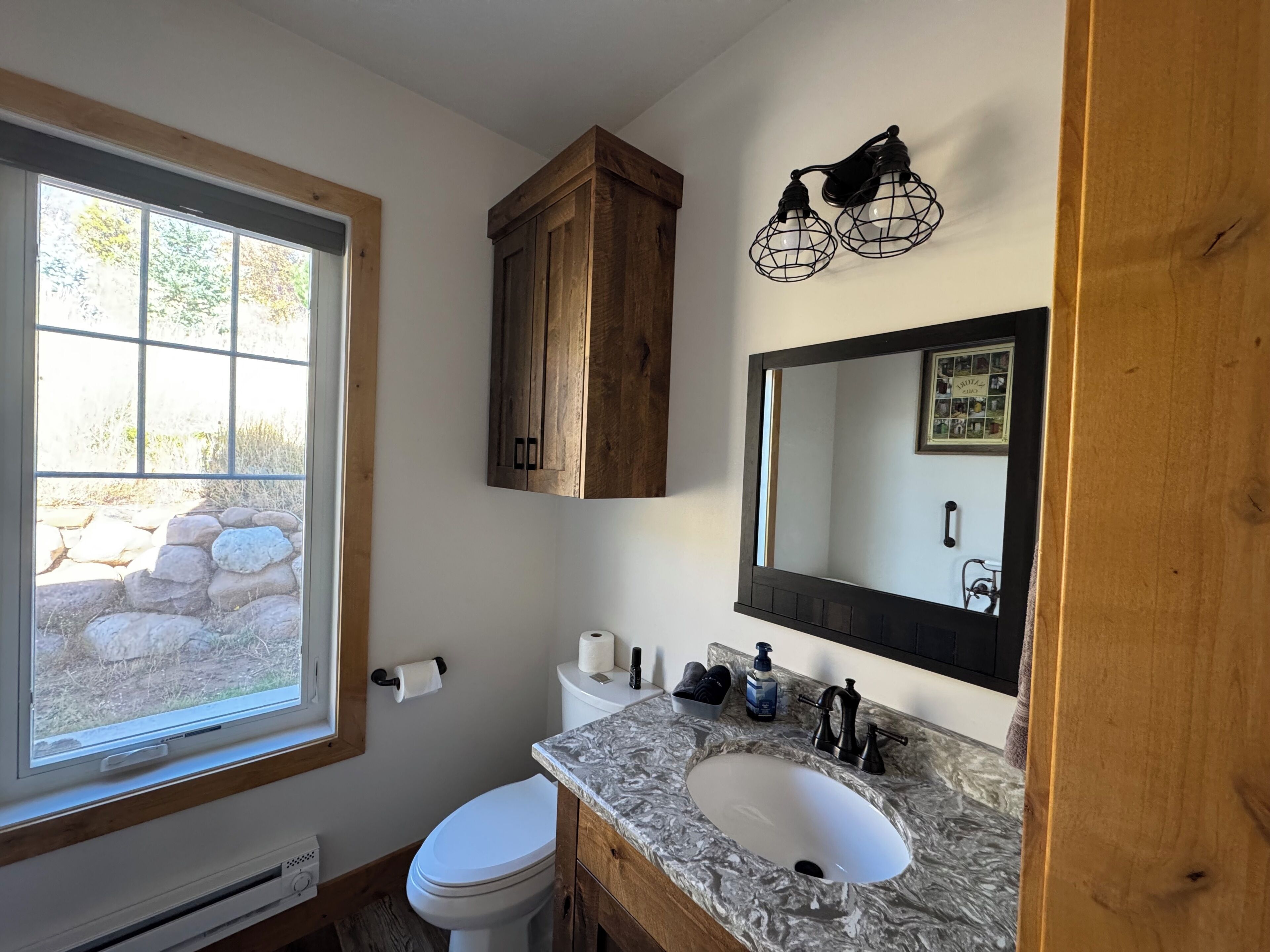 A bathroom with a window, a toilet, a sink with a marble countertop, and wooden cabinets.