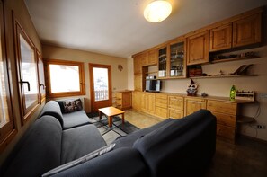 Una sala acogedora con un sofá y una mesita, armarios de madera y una zona de cocina con nevera y estanterías.