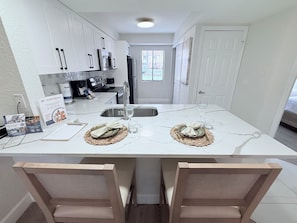 Cocina moderna con gabinetes blancos, electrodomésticos de acero inoxidable y encimera de mármol. Hay dos sillas de madera en la isla, y una ventana con persianas en el fondo.